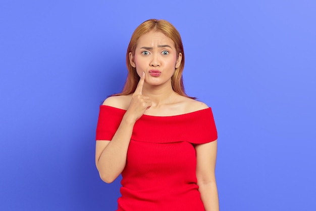 Portrait of innocent Asian woman wearing red dress looking forward with finger on her chin isolated on purple background