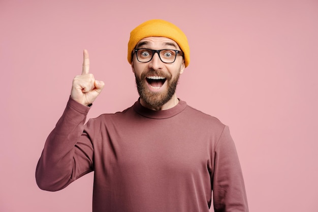 Foto ritratto di un uomo brunetto ispirato con la barba in cappello giallo e gli occhiali che puntano il dito verso l'alto