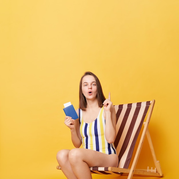 Portrait of inspired excited young brown haired woman wearing striped swimsuit holding document and ticket pointing finger up sitting on wooden chair isolated on yellow background