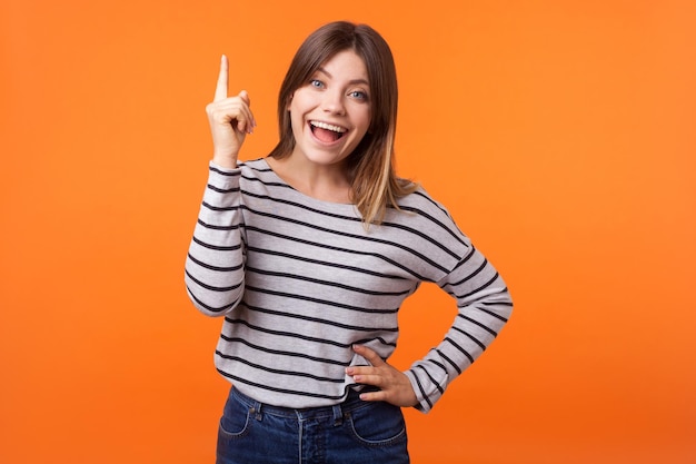 Portrait of inspired happy young woman with brown hair in long sleeve shirt standing with open mouth, pointing finger up to emphasize brilliant idea. indoor studio shot isolated on orange background