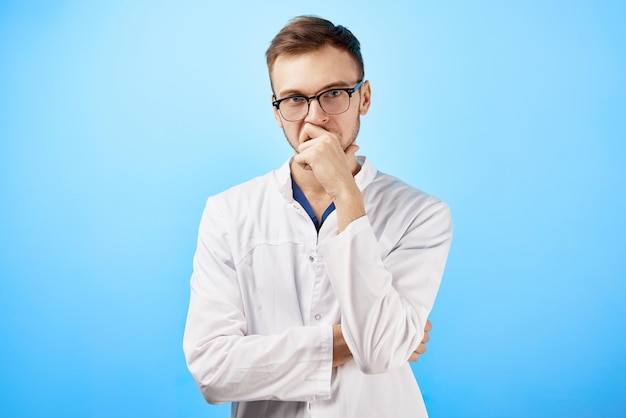 Portrait of intern doctor in white medical coat and glasses with a serious face expression isolated on blue wall background