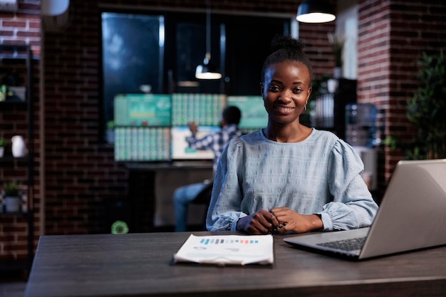 Portrait of investment company financial analyst sitting at desk in office while colleague working in background in front of multi monitor workstation. Stock market trader sitting in workspace.