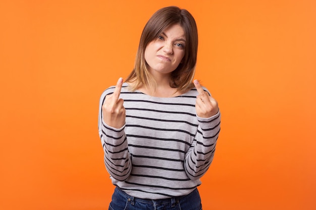 Портрет раздраженной агрессивной молодой женщины с каштановыми волосами в полосатой рубашке с длинным рукавом, стоящей со средними пальцами, оскорбительным жестом перед камерой. крытая студия снята на оранжевом фоне