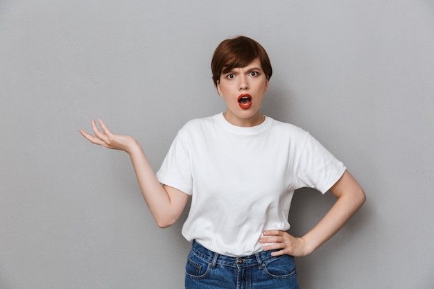 Portrait of irritated brunette woman wearing casual t-shirt frowning and gesturing hand isolated over gray wall