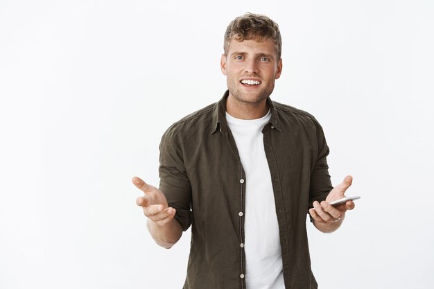 Portrait of irritated and confused good-looking caucasian man with blond hair raising hands in confusion and dismay holding smartphone, being questioned over gray wall