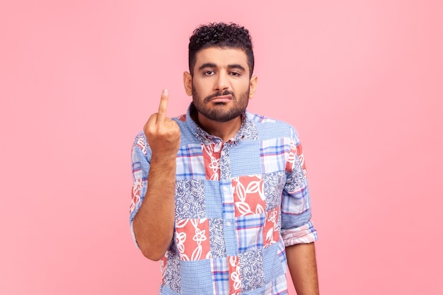 Portrait of irritated young adult man in blue casual style showing middle finger, impolite rude gesture of disrespect, hate and aggression in conflict. Indoor studio shot isolated on pink background.