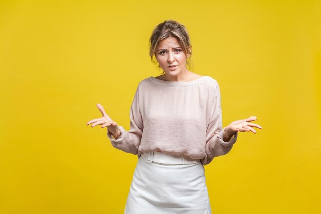 Foto ritratto di giovane donna irritata con capelli biondi in camicetta casual in piedi con le braccia alzate, guardando la fotocamera, chiedendo una risposta. tiro in studio indoor isolato su sfondo giallo