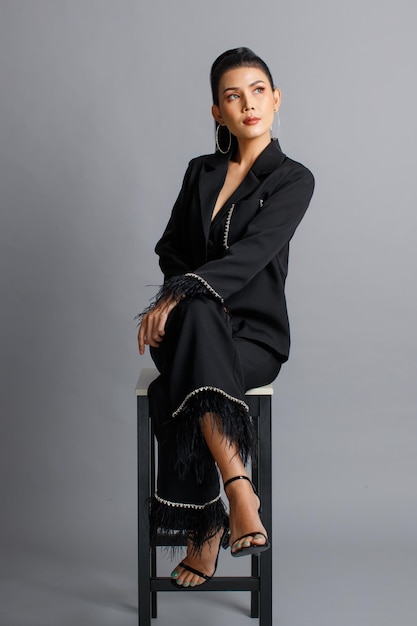 Portrait isolated cutout studio shot of asian female elegant\
fashion model in black casual fashionable fur suit and high heels\
sitting crossed legs on chair look at camera posing on gray\
background