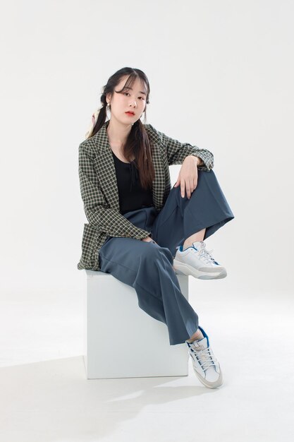 Portrait isolated cutout studio shot Asian young pretty female fashion model with pigtails braids hair in casual plaid suit crop top shirt sitting on square box posing look camera on white background