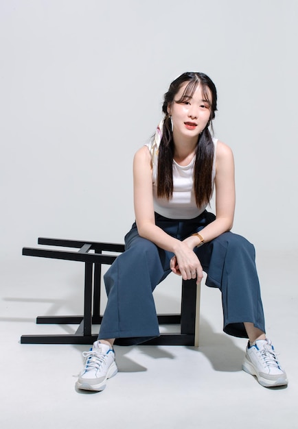 肖像画分離カットアウト スタジオ撮影アジアの若いきれいな女性ファッション モデルおさげ髪を三つ編みに髪をノースリーブ クロップ トップに座って背の高い椅子を横たわってポーズをとって白い背景にカメラを見て