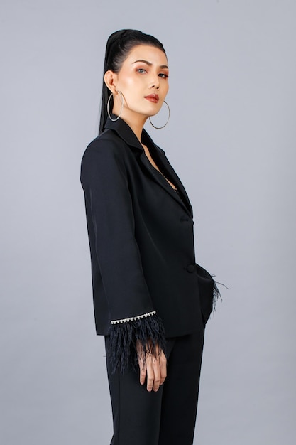 Portrait isolated cutout studio shot of millennial asian female\
elegant fashion model in black casual fashionable fur suit standing\
posing put hand in pants pocket look at camera on gray\
background