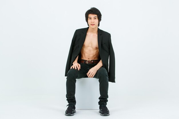 Portrait isolated studio shot Asian young handsome confident slim thin shirtless healthy athletic teen fashion male model in suit blazer jeans and sneakers sitting smiling posing on white background