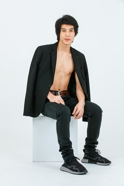 Portrait isolated studio shot Asian young handsome confident slim thin shirtless healthy athletic teen fashion male model in suit blazer jeans and sneakers sitting smiling posing on white background