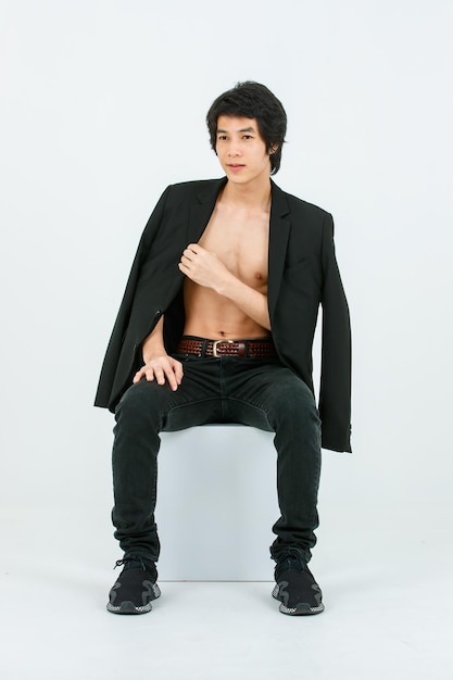 Portrait isolated studio shot Asian young handsome confident slim thin shirtless healthy athletic teen fashion male model in suit blazer jeans and sneakers sitting smiling posing on white background