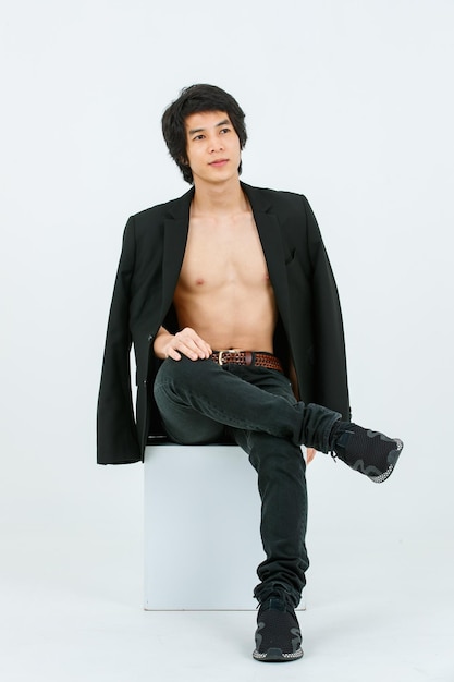 Portrait isolated studio shot Asian young handsome confident slim thin shirtless healthy athletic teen fashion male model in suit blazer jeans and sneakers sitting smiling posing on white background