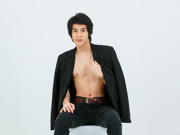 Portrait isolated studio shot Asian young handsome confident slim thin shirtless healthy athletic teen fashion male model in suit blazer jeans and sneakers sitting smiling posing on white background