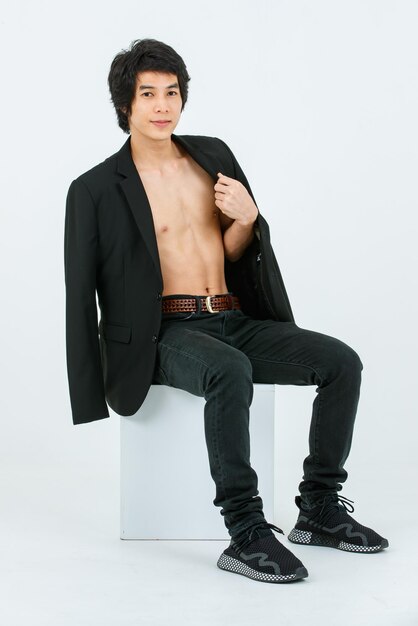 Portrait isolated studio shot Asian young handsome confident slim thin shirtless healthy athletic teen fashion male model in suit blazer jeans and sneakers sitting smiling posing on white background