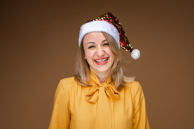 Portrait of jovial blonde Caucasian woman in yellow blouse wearing sparkling Santa hat