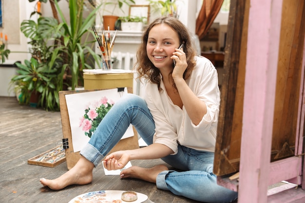 Portrait of joyful artistic woman sitting on floor and talking at cellphone while drawing picture on paper with paint palette in workshop or master class