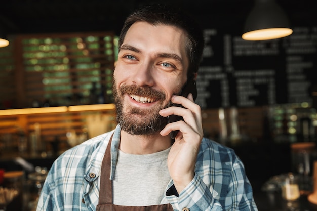 Portrait of joyful barista guy wearing apron smiling and talking on cellphone in street cafe or coffeehouse outdoor