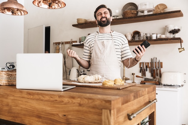 집에서 부엌에서 요리하고 집에서 만든 파스타를 만드는 동안 스마트폰을 사용하여 앞치마를 입은 즐거운 브루네트 남자의 초상화