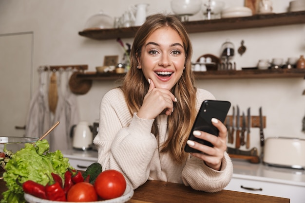 portrait of joyful caucasian woman wearing casual clothes using cellphone while cooking dinner in cozy kitchen