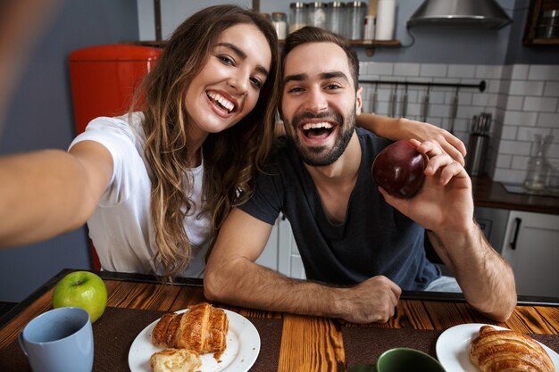 Portrait of joyful couple man and woman taking selfie photo on cell phone while having breakfast in kitchen at home