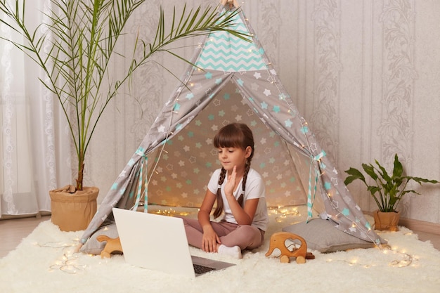Портрет радостной темноволосой маленькой девочки с косичками, лежащей в маленькой палатке с ноутбуком, которая машет рукой по видеозвонку и здоровается со своей подругой, смотрящей на экран устройства
