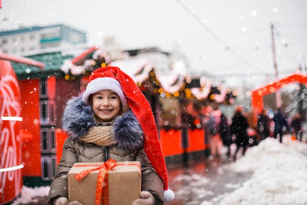 Portrait of joyful girl in Santa hat with gift box for Christmas on city street in winter with snow on festive market with decorations and fairy lights. Warm clothes, knitted scarf and fur. New year