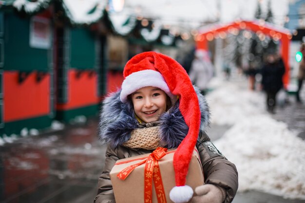 Portrait of joyful girl in Santa hat with gift box for Christmas on city street in winter with snow on festive market with decorations and fairy lights. Warm clothes, knitted scarf and fur. New year