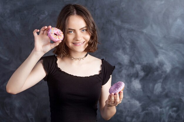 Portrait of joyful girl with donuts on black wall. Happy girl holding fresh donuts and smiling. Good mood, diet concept.