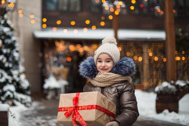 Portrait of joyful girl with a gift box for christmas on a city\
street in winter with snow on a festive market with decorations and\
fairy lights warm clothes knitted hat scarf and fur new year