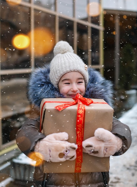 크리스마스 선물 상자가 있는 즐거운 소녀의 초상화는 장식과 조명이 있는 축제 시장에 눈이 내리는 겨울에 도시 거리에 있습니다. 따뜻한 옷, 니트 모자, 스카프 및 모피. 복사 공간