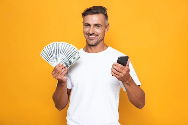 Portrait of joyful guy 30s in white t-shirt holding cell phone and bunch of money isolated on yellow