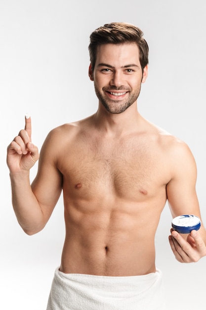 Portrait of joyful half-naked man in bath towel smiling and showing facial cream isolated on white