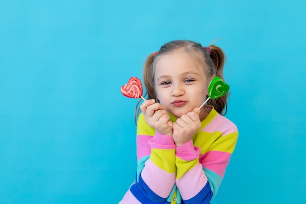 Portrait of a joyful little girl with lollipops in a striped jacket a large candy on a stick The concept of sweets and confectionery Blue background photo studio place for text