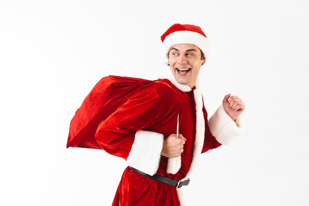 Portrait of joyful man 30s in santa claus costume and red hat running with gift bag over shoulder