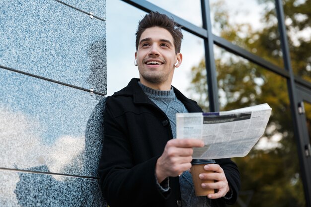 Foto ritratto di un gioioso uomo di 30 anni che indossa auricolari che legge il giornale e tiene in mano caffè da asporto mentre si trova di fronte a un edificio di vetro glass