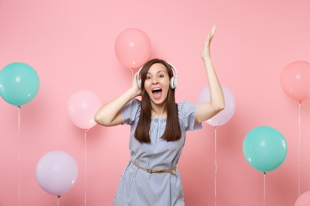 Portrait of joyful pretty young woman with opened mouth with headphones wearing blue dress listening music raising hands on pink background with colorful air balloons. Birthday holiday party concept.