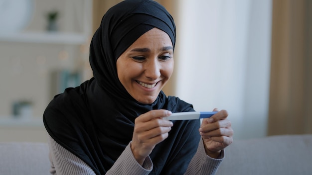 Portrait joyful surprised girl in hijab holding pregnancy test sit in living room excited