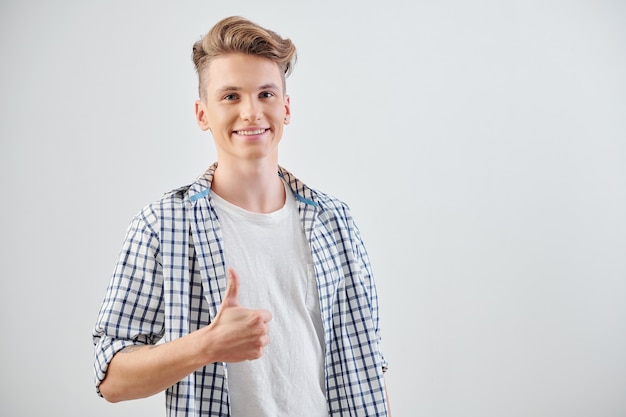 Portrait of joyful teenage boy with toothy smile showing thumbs-up and looking