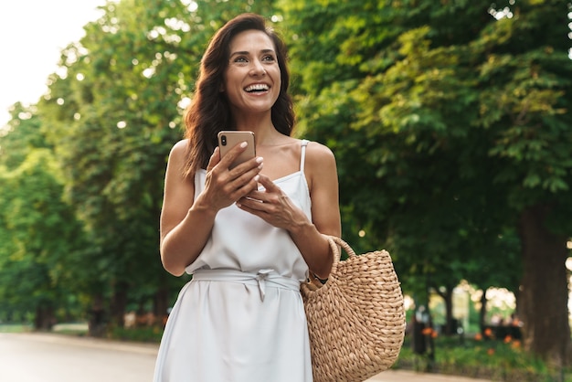 Portrait of joyful woman in summer dress smiling and holding cellphone while walking through green boulevard