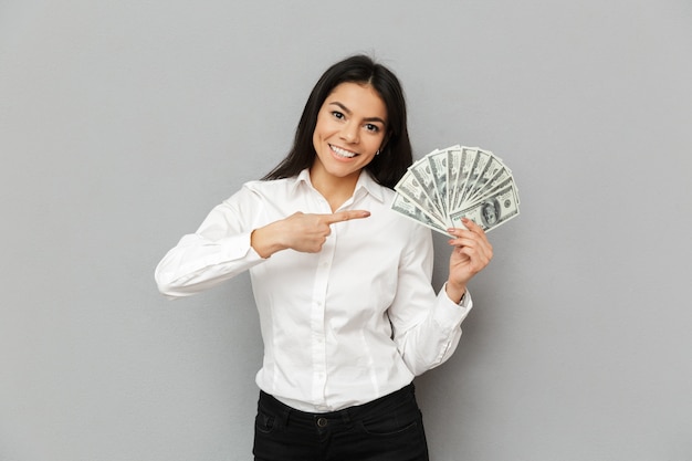 Портрет радостной женщины с длинными каштановыми волосами в офисной одежде, улыбающейся и указывающей пальцем на много долларов в руке, изолированной над серой стеной
