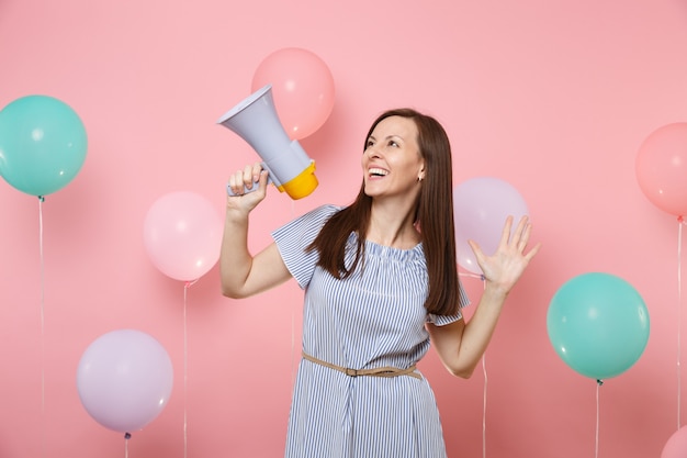 Portrait of joyful young attaractive woman wearing blue dress holding megaphone spreading hands on pink background with colorful air baloons. Birthday holiday party, people sincere emotions concept.