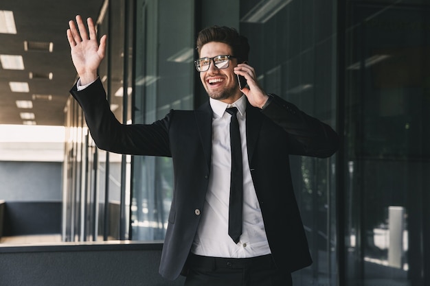 Portrait of joyful young businessman dressed in formal suit standing outside glass building, and talking on mobile phone