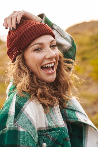 Portrait of joyful young caucasian woman wearing hat and plaid shirt smiling while walking outdoors