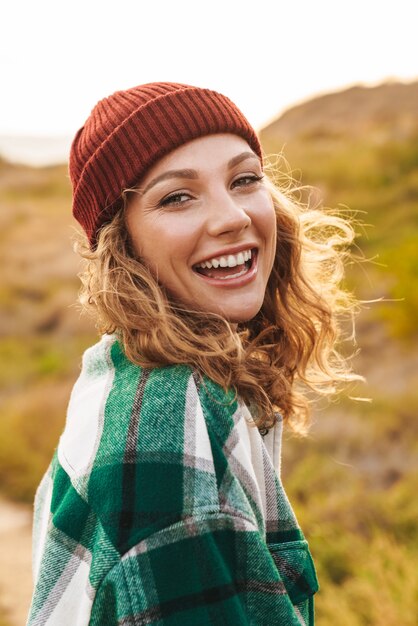 Portrait of joyful young caucasian woman wearing hat and plaid shirt smiling while walking outdoors