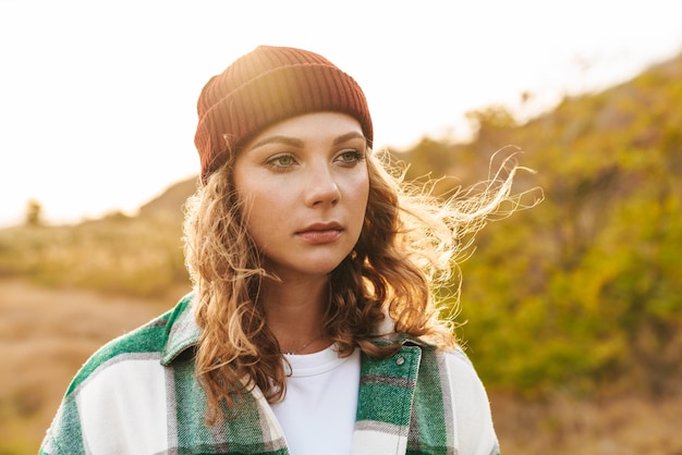 Portrait of joyful young caucasian woman wearing hat and plaid shirt smiling while walking outdoors