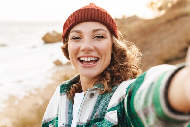 Portrait of joyful young caucasian woman wearing hat and plaid shirt taking selfie Portrait while walking outdoors