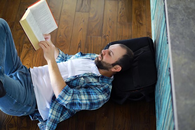 Ritratto di giovane gioioso che legge un libro mentre è seduto sul pavimento nel suo soggiorno studente che tiene e legge il libro